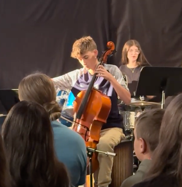 A young boy plays a cello with a band; another band member plays drums behind him. An audience watches.