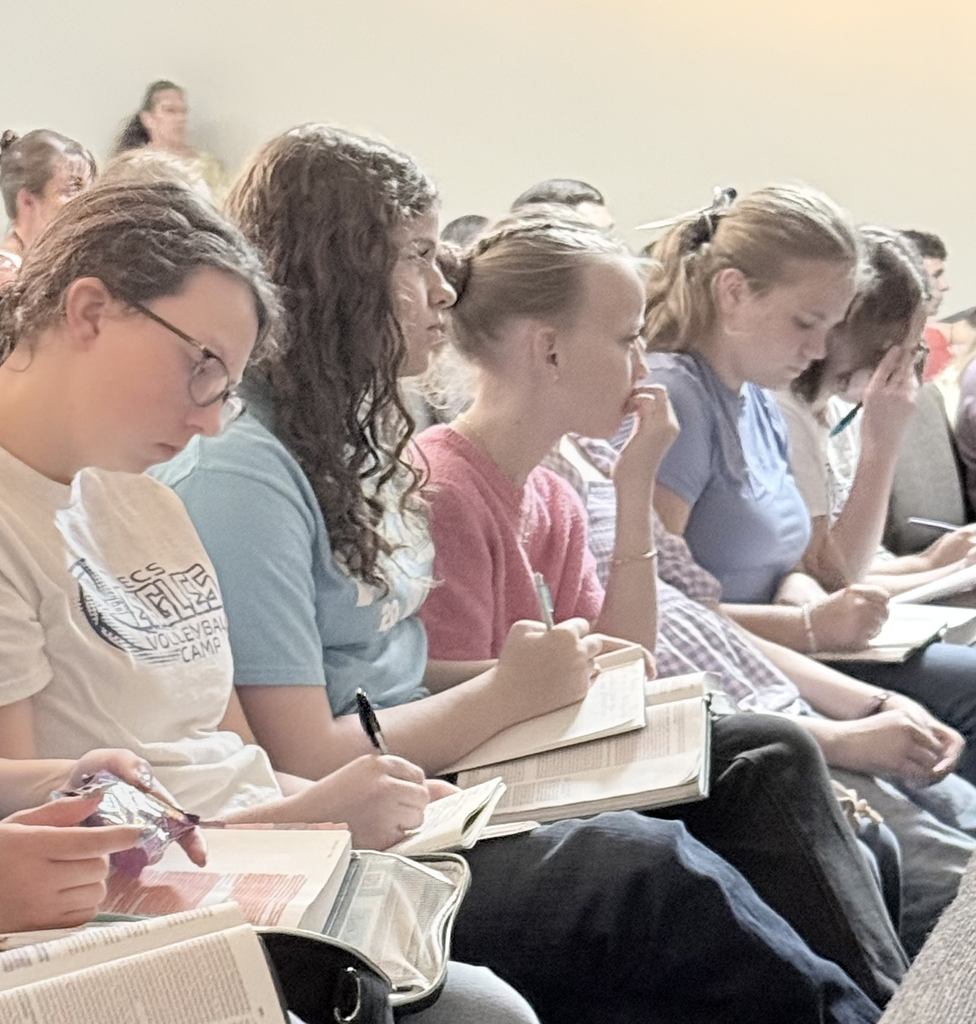 A classroom of students taking notes in a large book with pens. They are sitting in rows.