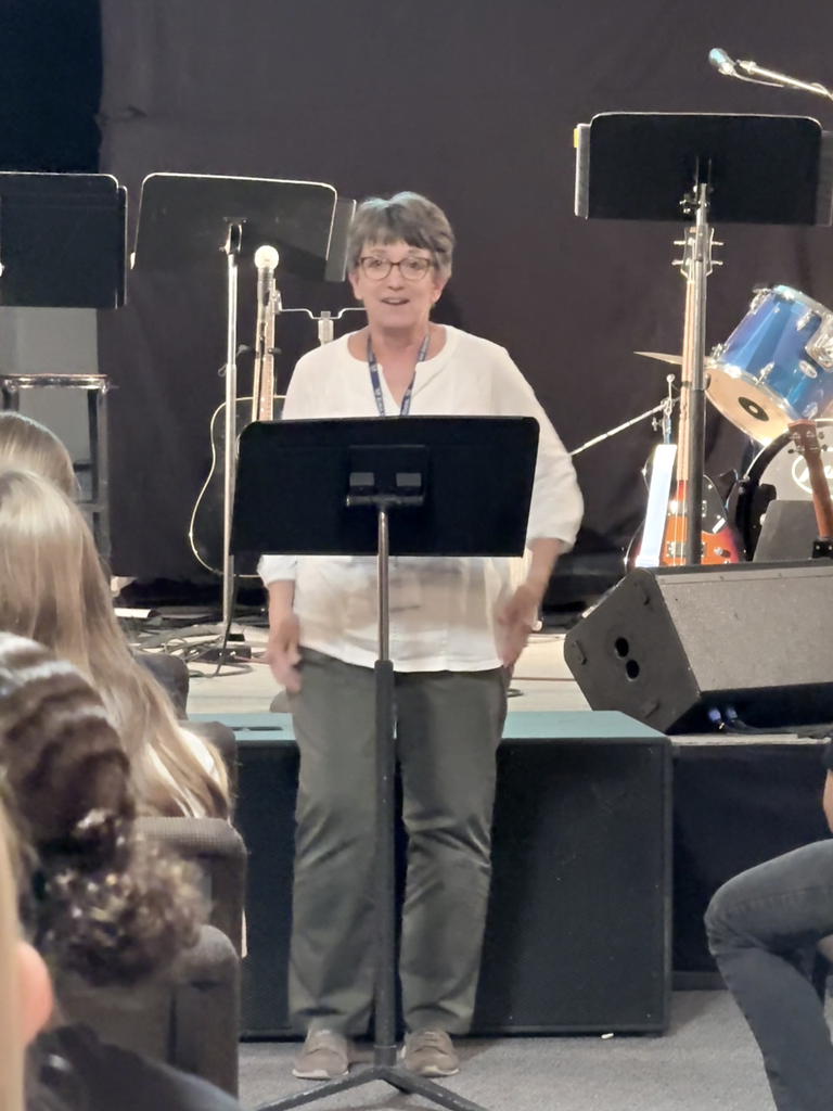 A woman stands behind a music stand in front of a crowd. Behind her, various musical instruments are arranged.