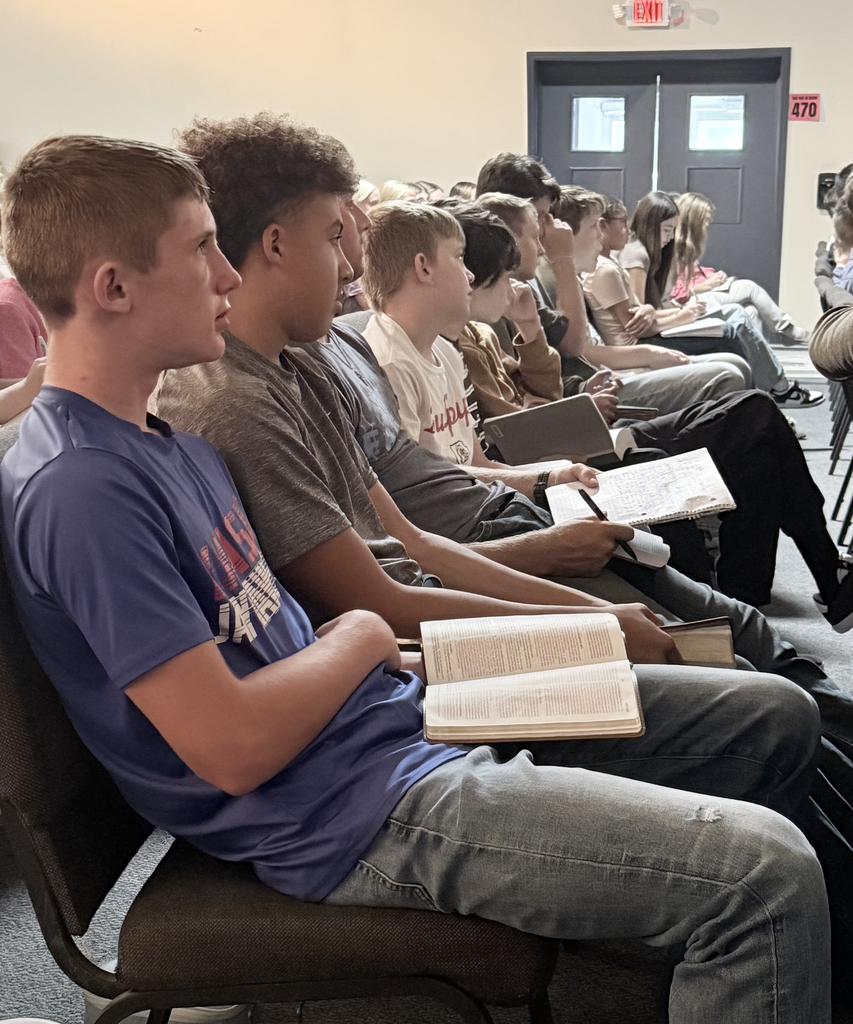 Several individuals seated in a row, looking ahead. One holds an open book. A double door is at the back.