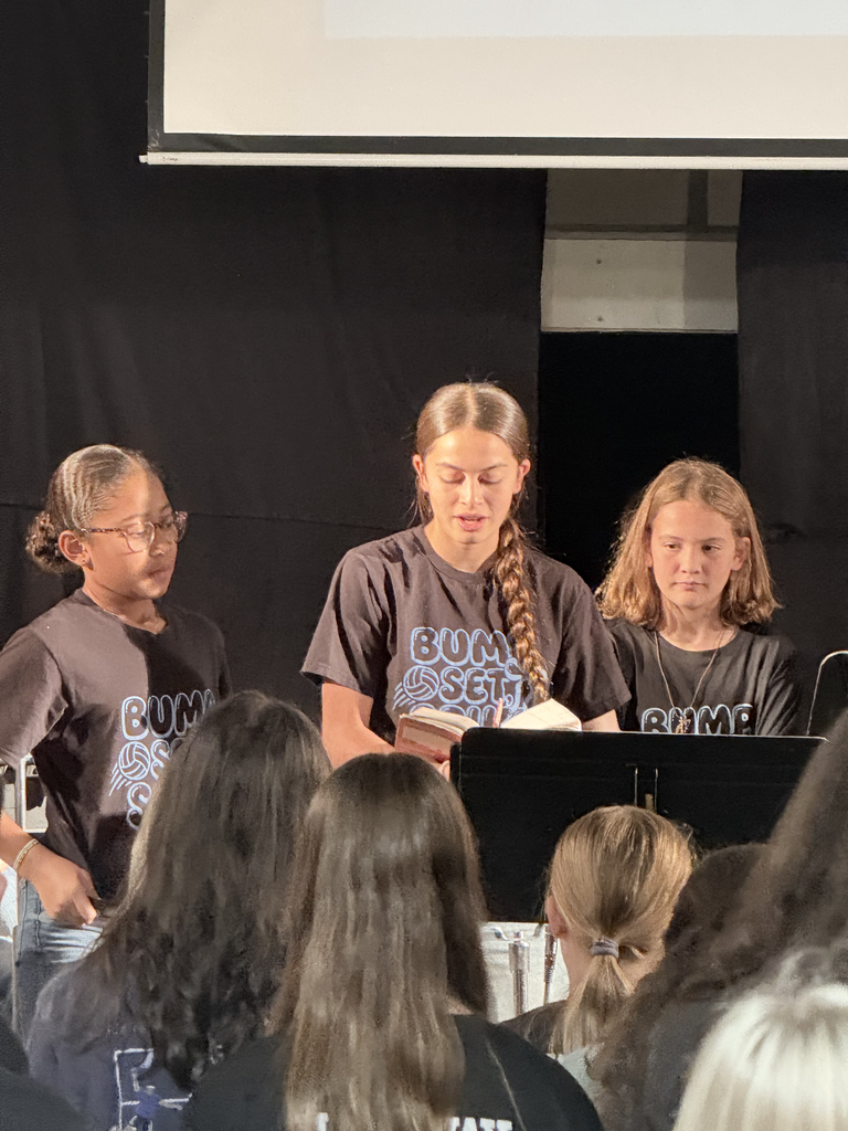 Three girls in matching shirts, one reading from a book, in a darkened room. Audience members are visible in the foreground.