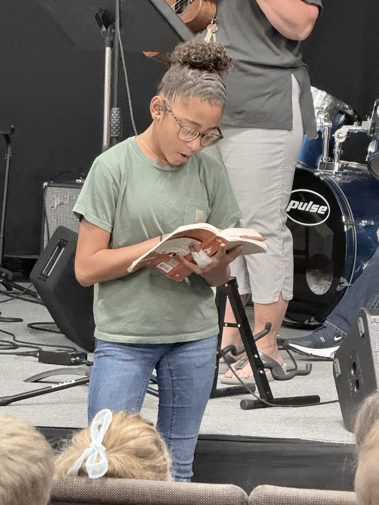 Girl reading a book on a stage, wearing glasses. Behind her, another person stands, holding a musical instrument.