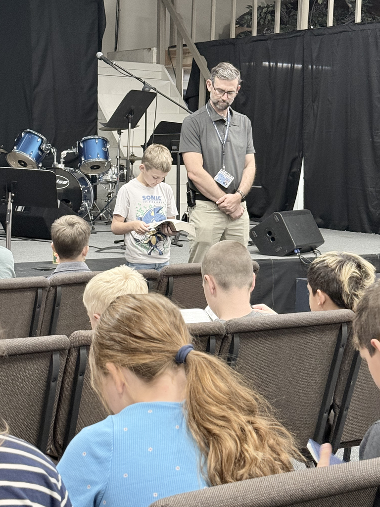A boy on a stage holds a book and reads aloud, while a man stands beside him. Children sit in front of them in chairs.