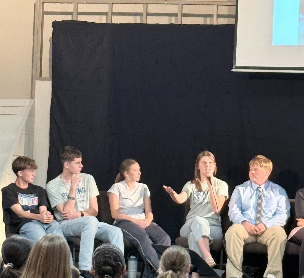 Several people sit on stage in front of a dark backdrop. A projection screen is visible above them.