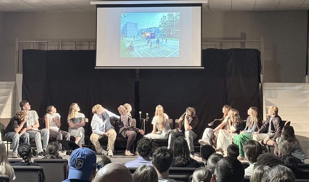 People seated on a stage, facing an audience, with a projector screen displaying an image above them.
