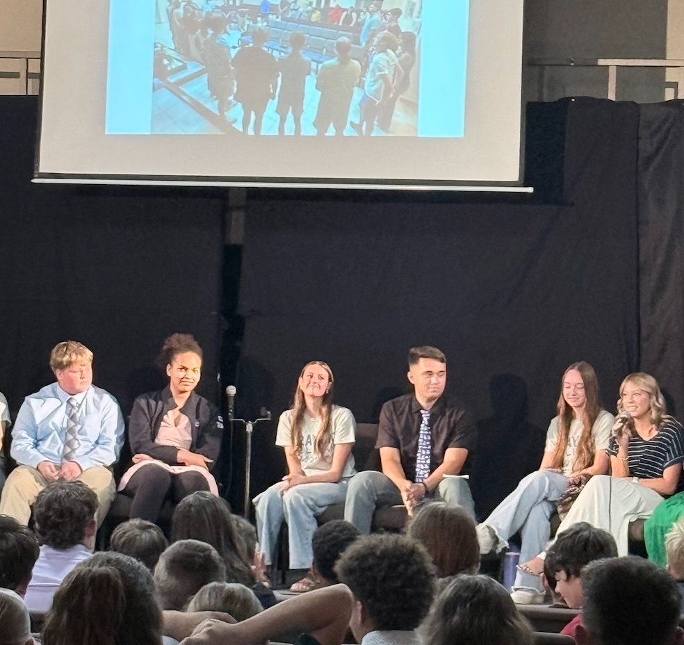 A group of people are seated on chairs at the front of a room. A projector screen displays a blurred image of a crowd.