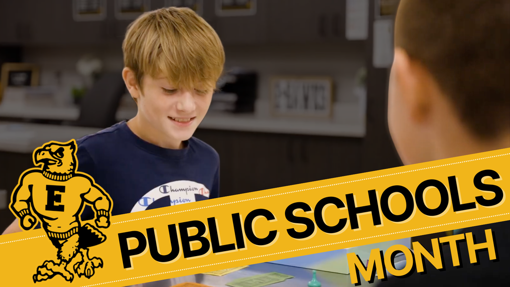 Student smiling at a table with classroom materials, overlaid with yellow banner reading “Public Schools Month” and school mascot logo.