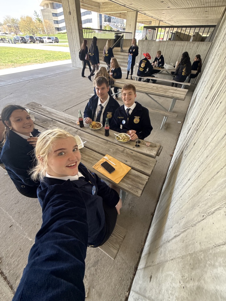 FFA students gathered at outdoor picnic tables under a concrete overhang, eating and chatting; one student takes a selfie with classmates in the background.