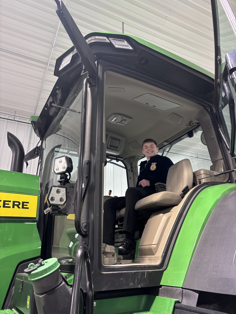 Teen in an FFA jacket sits in the driver’s seat of a large green tractor, smiling inside a garage-like space.