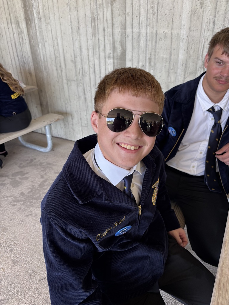 Close-up of a smiling teen in an FFA jacket wearing reflective sunglasses, seated beside another student at a picnic table.