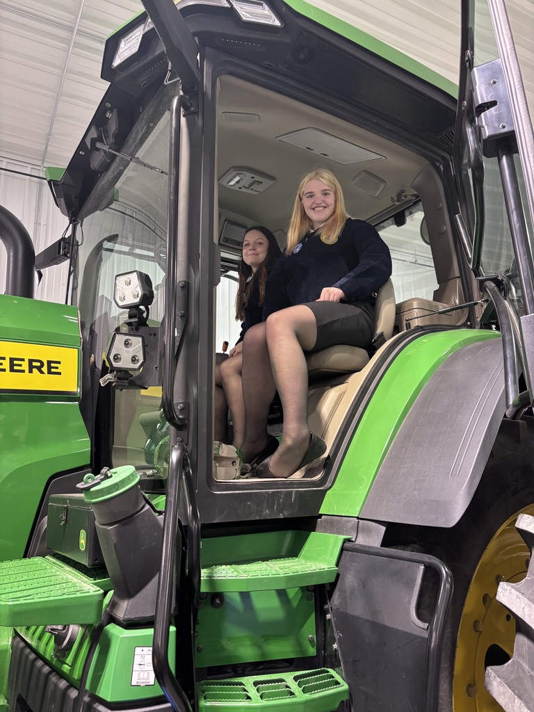 Two students in FFA jackets sit inside a large green tractor cab, smiling, one in the driver’s seat and one beside her.