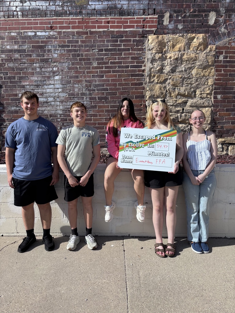 Group of five teens standing outdoors by a brick wall; one holds a sign reading “We Escaped From No Escape Iowa in 54.47 minutes, Emmetsburg FFA.”