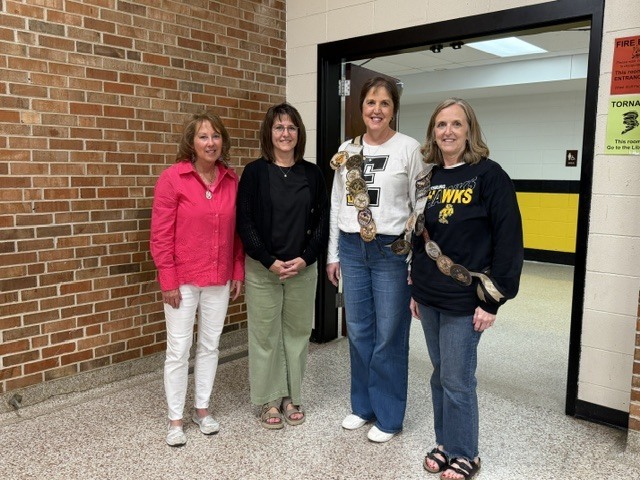Four staff members stand together in a school hallway, smiling for a group photo near a brick wall and classroom entrance.