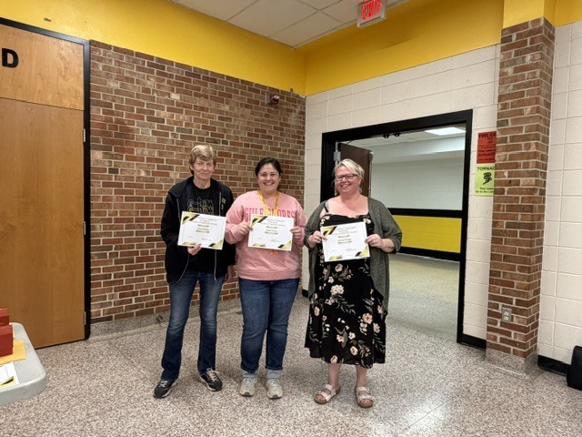 Three staff members stand in a school hallway holding certificates and smiling during a recognition or award presentation.
