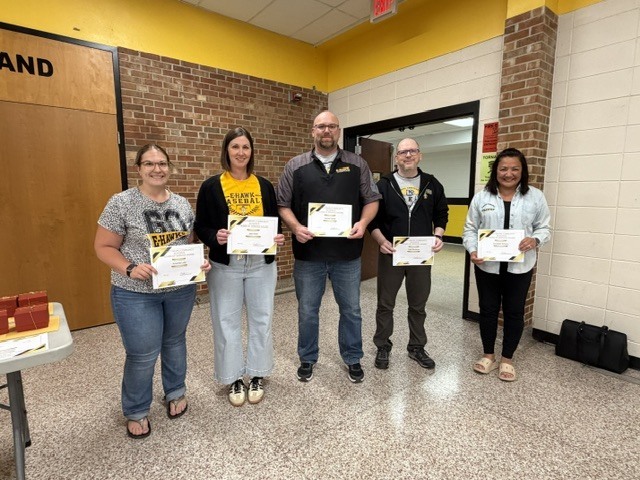 Five staff members stand in a school hallway holding certificates and smiling during an employee recognition or award presentation.