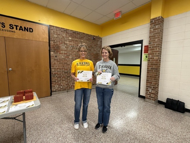 Two staff members stand in a school hallway holding certificates and smiling during an employee recognition or award presentation.