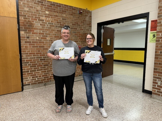 Two staff members stand in a school hallway holding certificates and smiling during an employee recognition or award presentation.