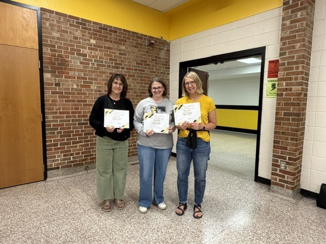 Three staff members stand in a school hallway holding certificates and smiling during an employee recognition or award presentation.