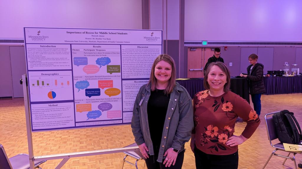 Two women smile beside a research poster titled “Importance of Recess for Middle School Students” at an academic event.