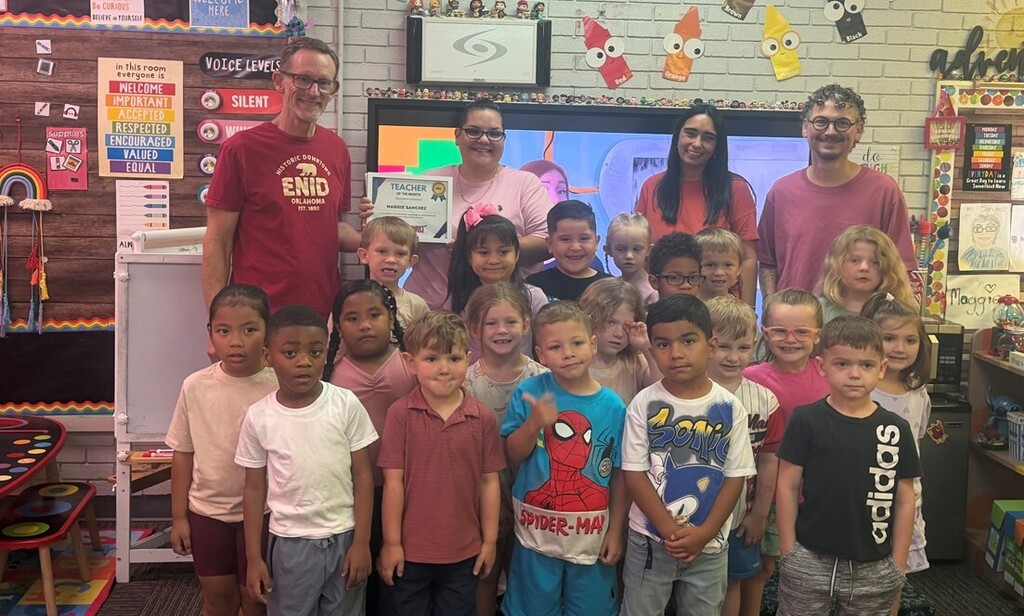 Four adults and a group of young elementary students pose in a decorated classroom while holding a "Teacher of the Month" award certificate for Maggie Sanchez.