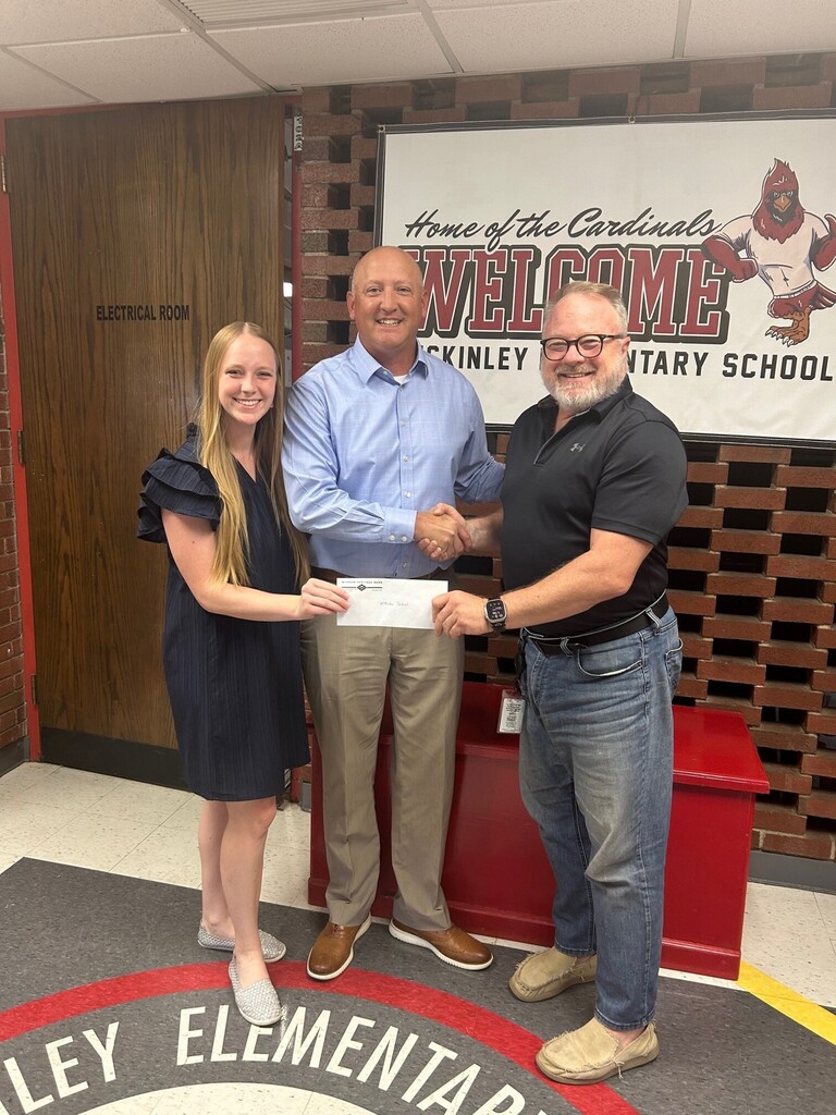 Three people pose inside McKinley Elementary School while shaking hands and holding a donation envelope in front of a school banner.