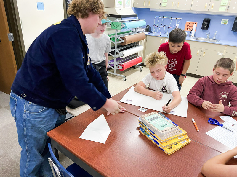 FFA high school student leans over to help middle school students working at a desk on a project