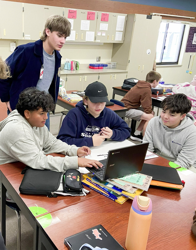 high school student looking over the shoulder of middle school students in a group seated at a desk working together on a Chromebook