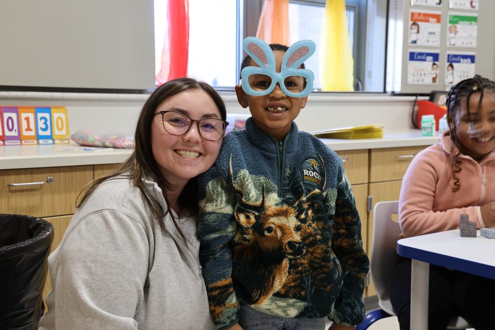 A teacher smiling next to a student who is wearing glasses in the shape of rabbit ears.