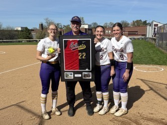 Erie High School GIrl's Varsity Softball Head Coach Ron Niebauer stands with framed jerseys honoring his 50 years of coaching in Erie's Public Schools. Three softball players stand  beside him.
