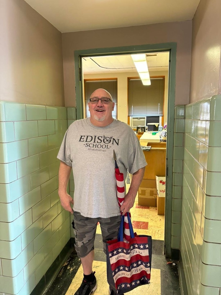A man is posing in front of the library door by holding American flag and a bag.