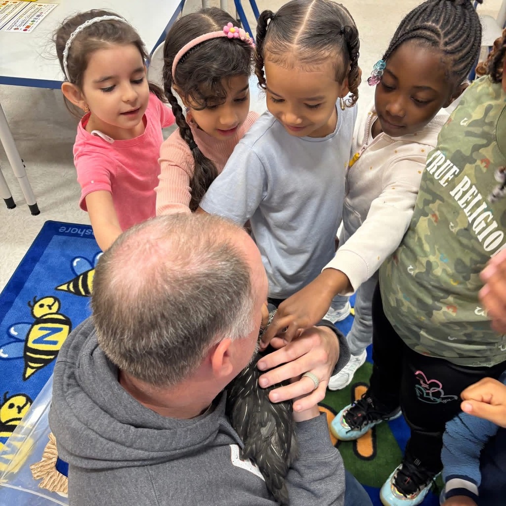 Children gather around a man holding a small chicken, reaching out to touch it in a classroom. 