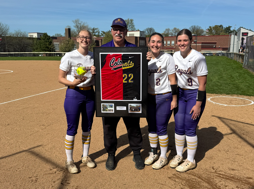 Coach Ron Niebauer smiling with the Erie High Royals Girls Softball team and holding a frame with half of a Strong Vincent jersey and half of an Erie High jersey. 