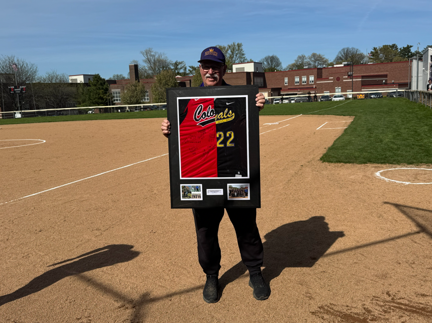 Coach Ron Niebauer smiling and holding a frame with half of a Strong Vincent jersey and half of an Erie High jersey. .