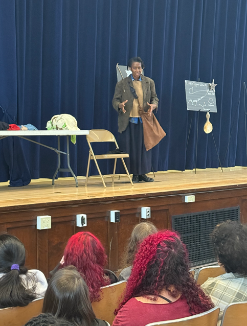 An actor dressed as Harriet Tubman is speaking to a room of students. 