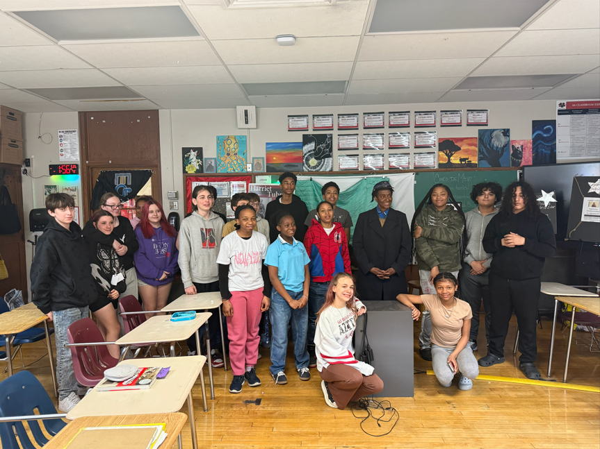 An actor dressed as Harriet Tubman is smiling with students in a classroom. 