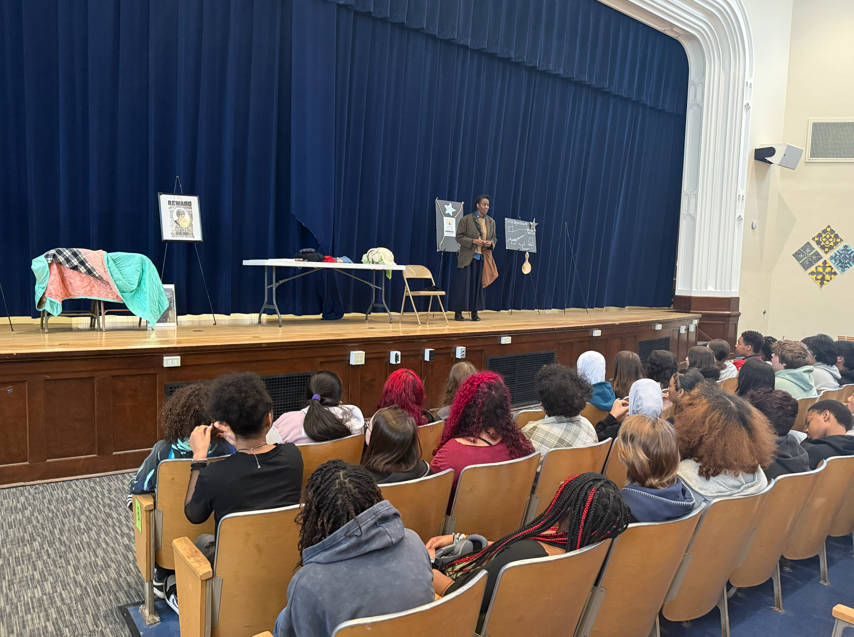 An actor dressed as Harriet Tubman is speaking to a room of students. 