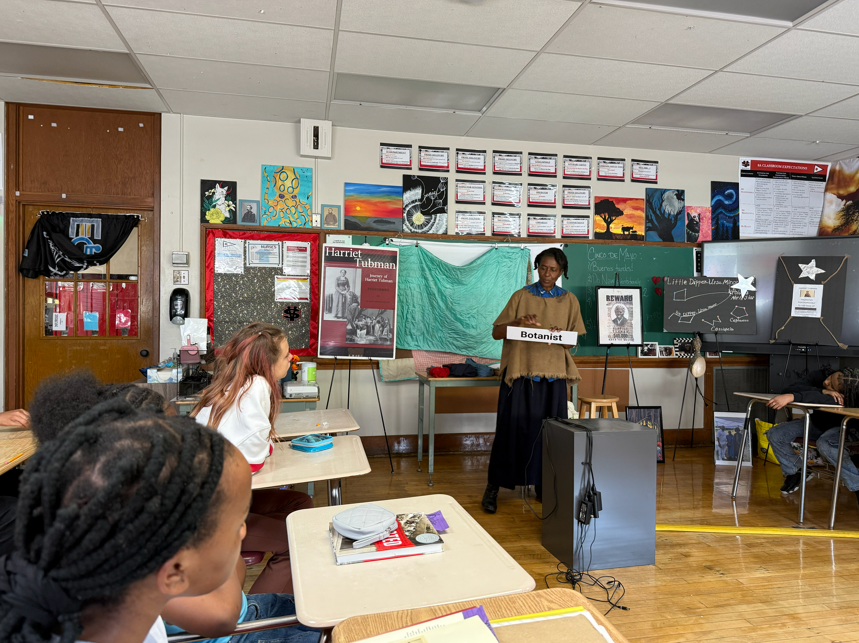 An actor dressed as Harriet Tubman is speaking to a room of students. 