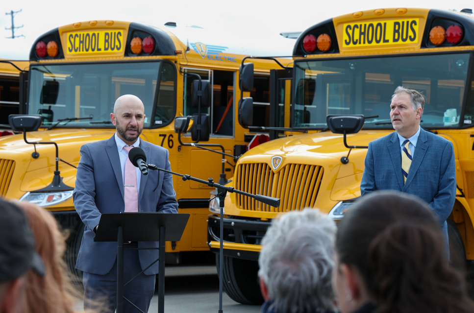 A man wearing a gray suit is talking into a microphone in front of a yellow school bus.