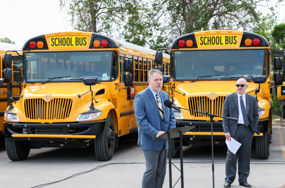 A man wearing a gray suit is talking into a microphone in front of a yellow school bus.