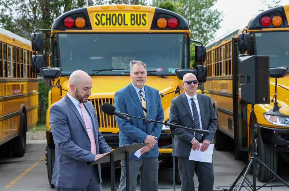 Three men standing in front of new school buses while one of them speaks into a microphone.