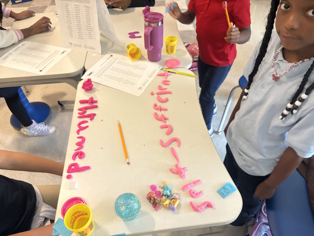 Student smiling. On the desk in front of him is a list of spelling words and the word "self-sufficient" spelled out using play-doh.
