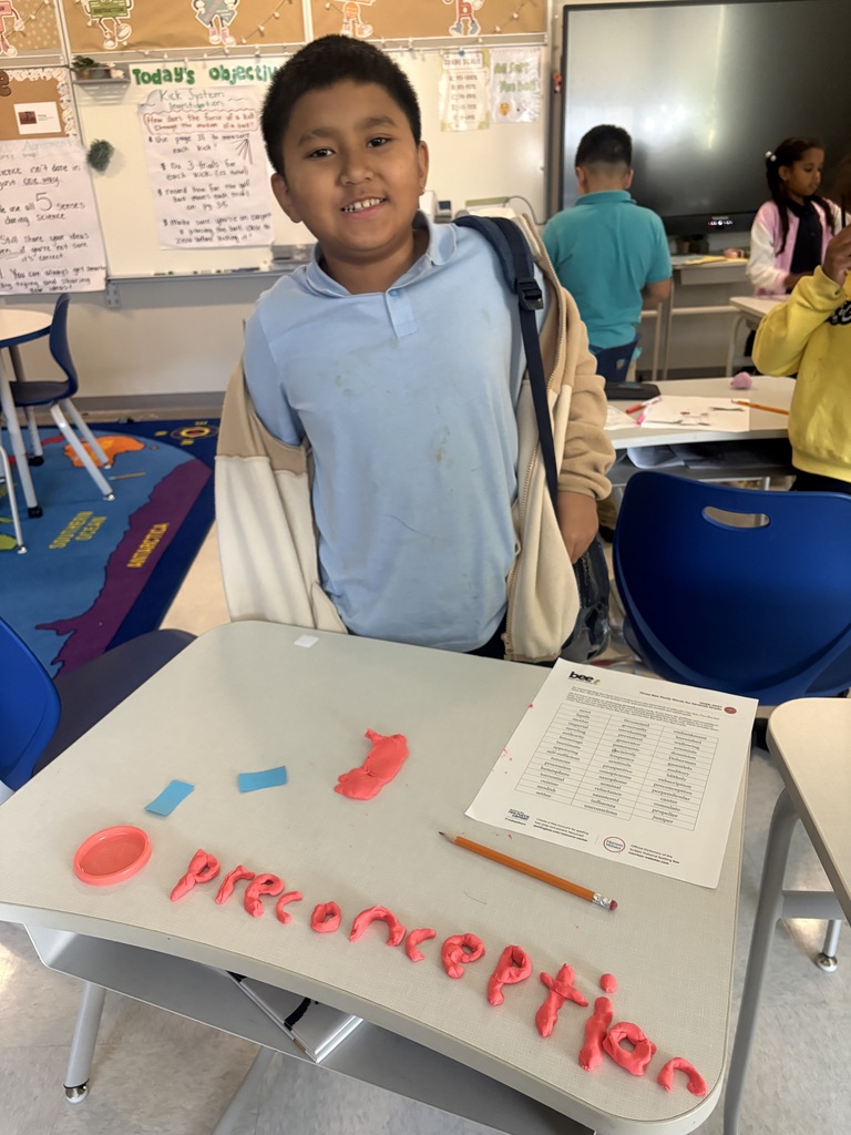 Student smiling. On the desk in front of him is a list of spelling words and the word "preconception" spelled out using play-doh.