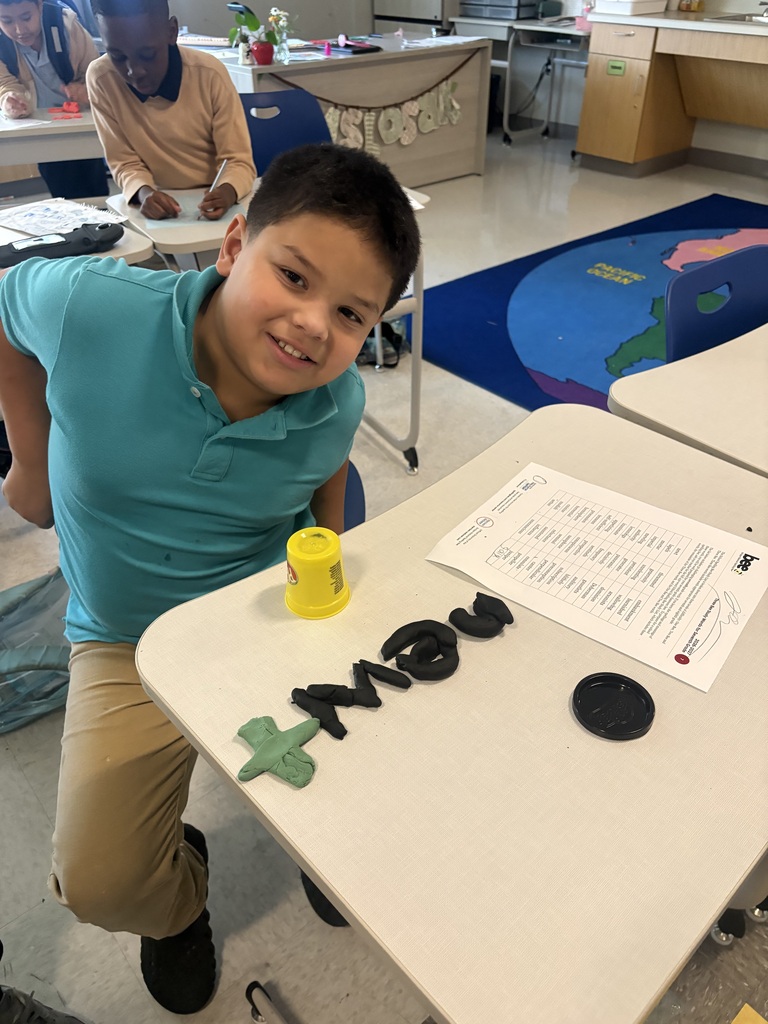 Student smiling. On the desk in front of him is a list of spelling words and the word "newt" spelled out using play-doh.