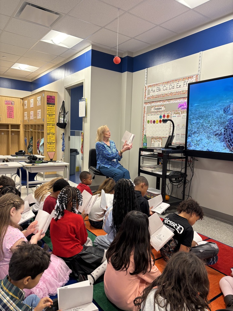 Guest reader holding up the book she is reading, while students on the floor look through their copies.