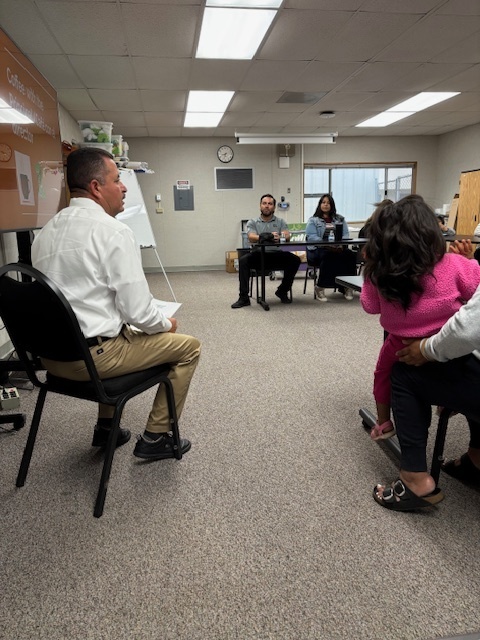 Principal Lopez is sitting on a black chair and provides information about school events to parents attending coffee with the principal