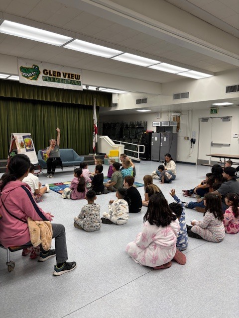 Mrs. Kim lifts up her arm while reading a story to one parent and 22 students inside the multipurpose room
