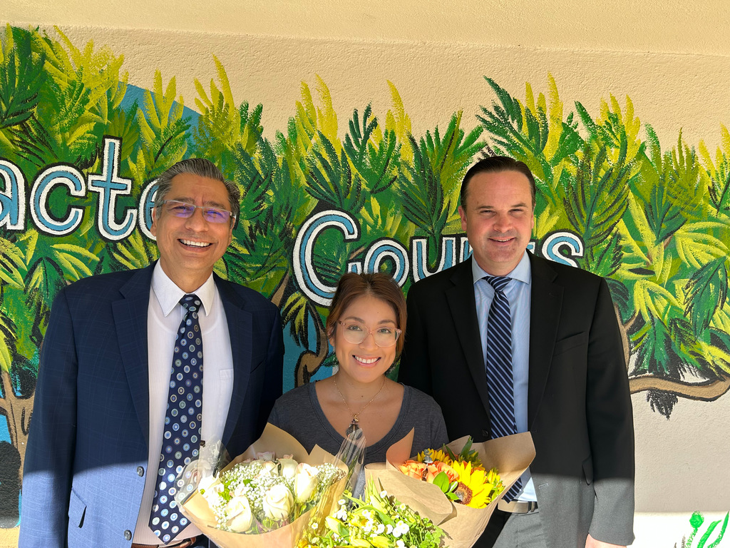Three people posing in front of a leaf-themed mural, with one holding a bouquet of flowers.