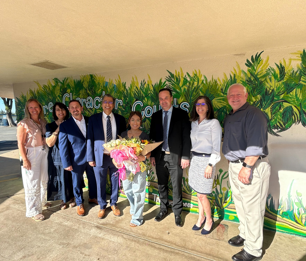 A group of eight people stands in front of a mural with greenery and the words "Character Counts," one person holding a bouquet.