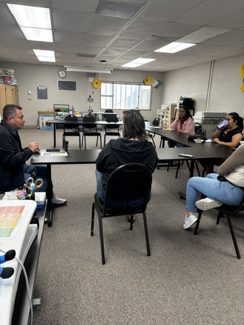 Principal Lopez is sitting on a chair and is speaking to four parents inside room 21. 