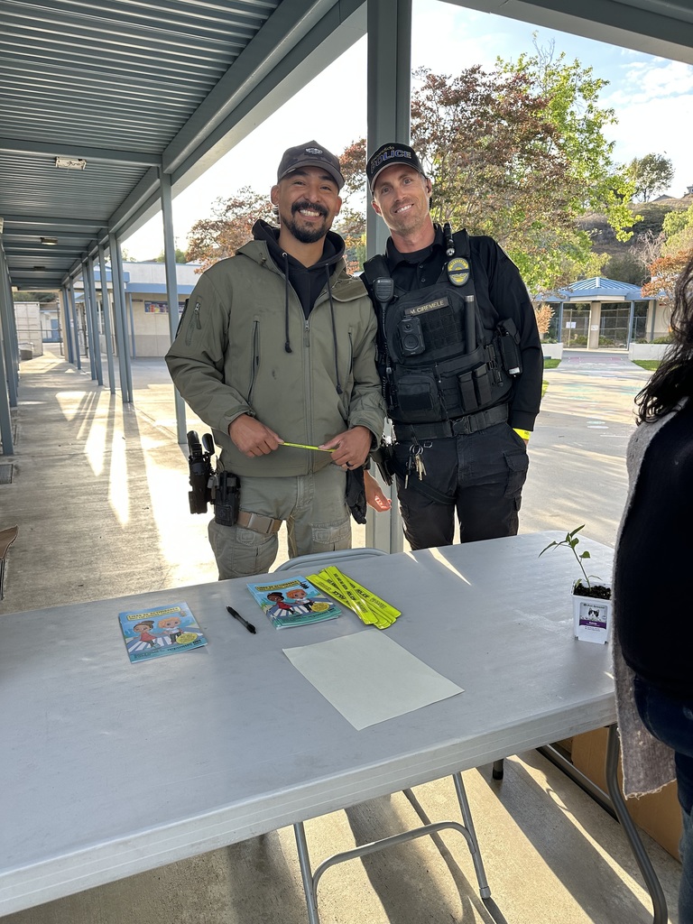 "Two law enforcement officers smile at the camera while standing at an information table at an outdoor school community event."